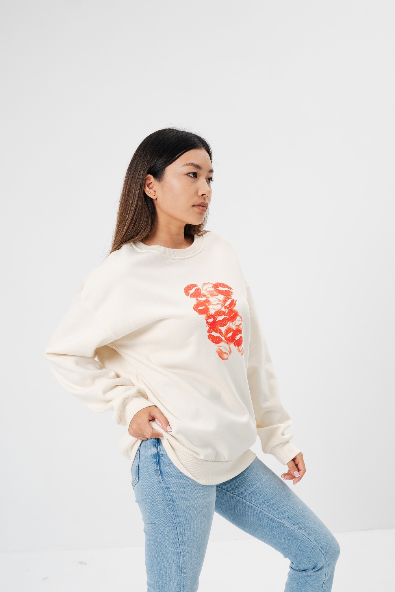 Jasna bluza z nadrukiem ust na modelce, w jasnych jeansach. Ujęcie z profilu, na białym tle. Widoczna biżuteria i delikatny makijaż.