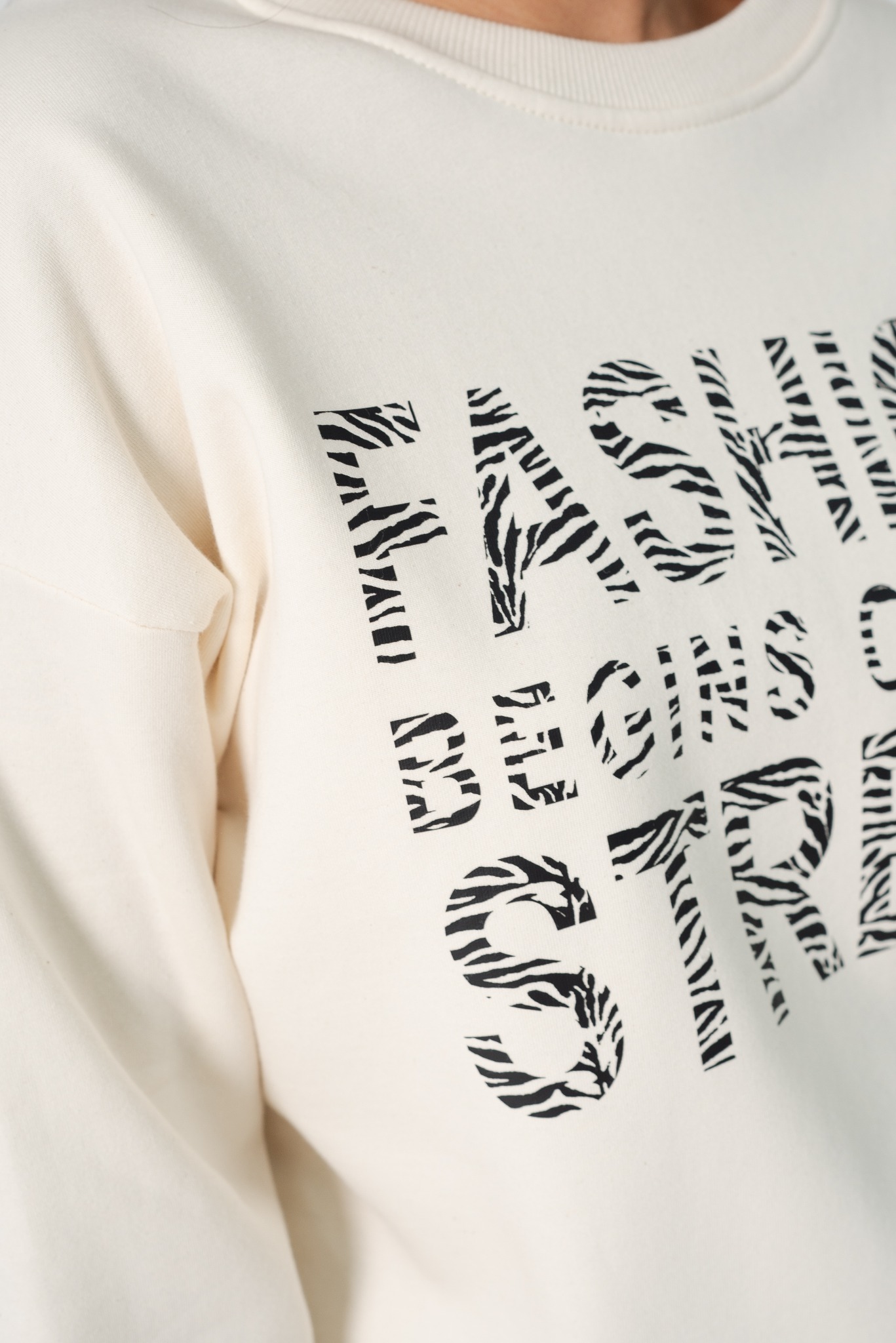 Kremowa bluza z napisem 'Fashion Begins Striking' wypełnionym wzorem zebry, widok z bliska na klatkę piersiową i ramię.