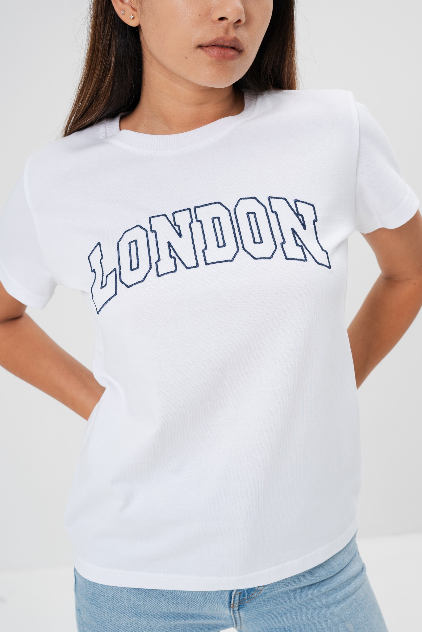 Biała koszulka z granatowym napisem 'LONDON' na modelce, ręce w kieszeniach, jasne jeansy, widoczne kolczyki w uszach.