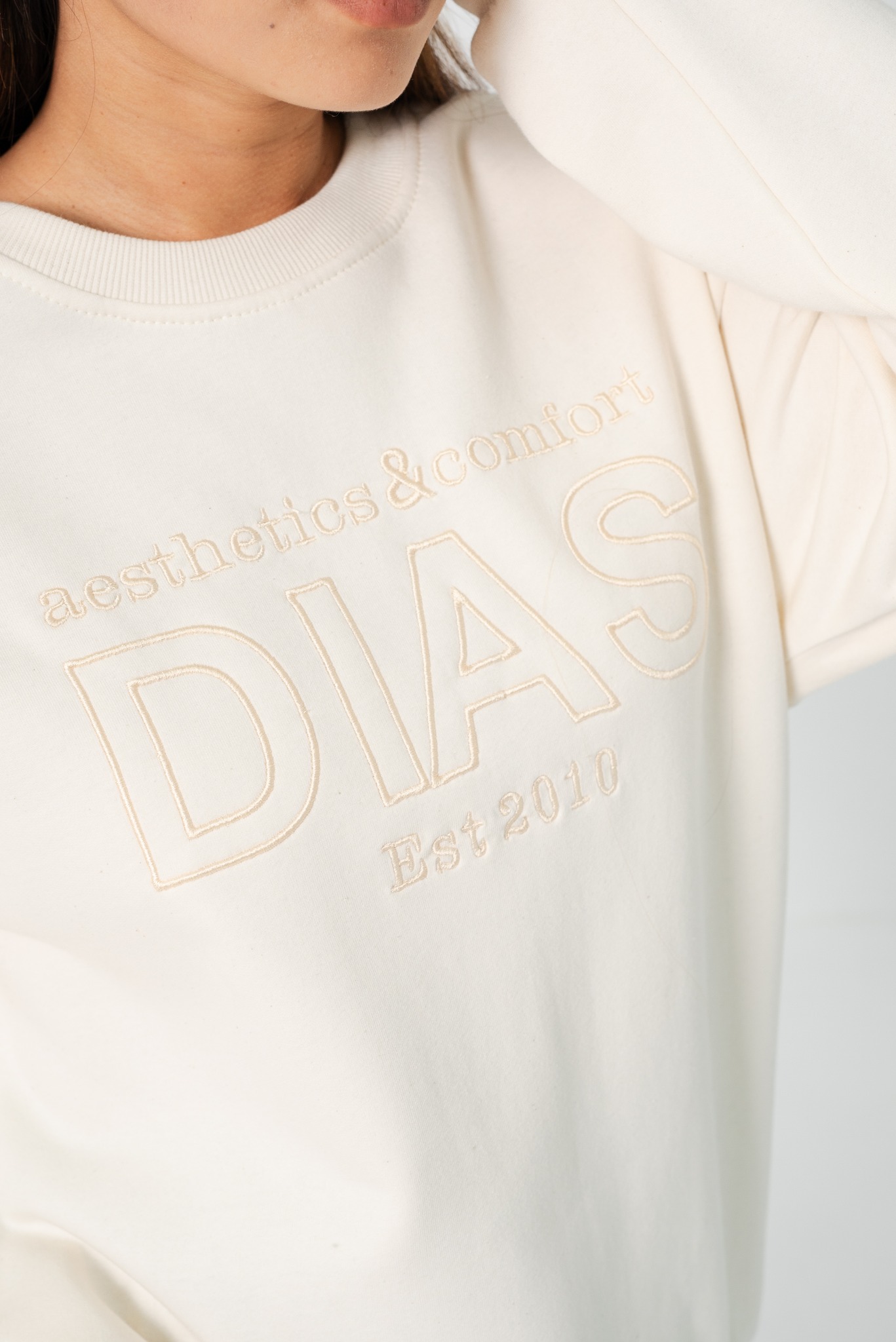 Kremowa bluza z beżowym, delikatnym haftem 'DIAS aesthetics&comfort Est.2010', widoczna na osobie od szyi w dół. Minimalistyczny design.