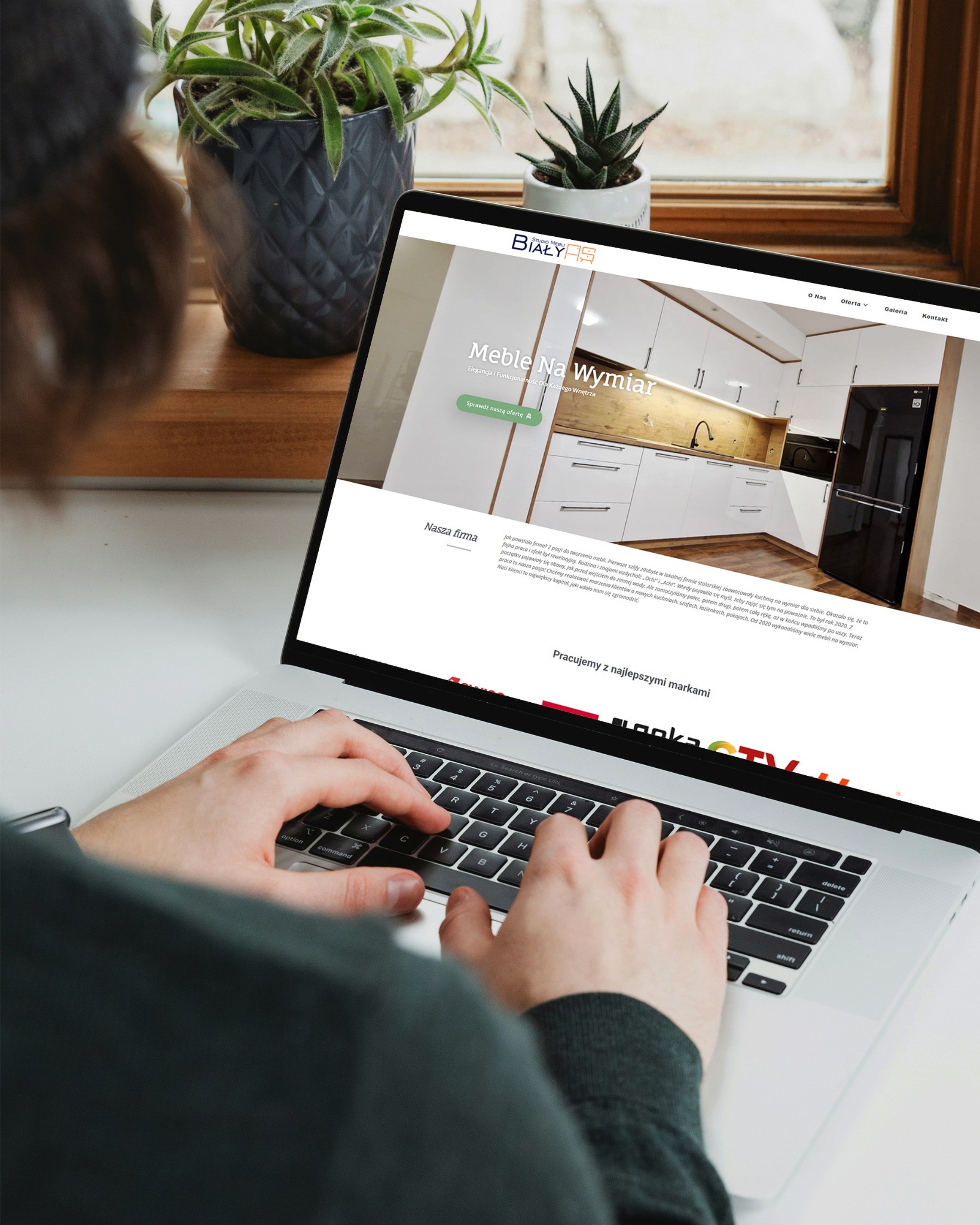 Osoba pisze na laptopie, na ekranie strona internetowa studia mebli na wymiar 'Białys' z aranżacją kuchni. Za laptopem rośliny doniczkowe i okno.