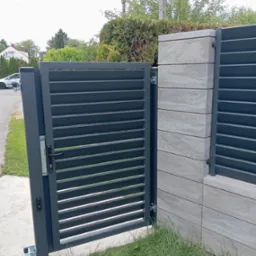 Nowoczesna, otwarta brama lamelowa w grafitowym kolorze, zamontowana na betonowym słupku z widocznymi poziomymi warstwami. Brama częściowo otwarta, z widocznym zamkiem.