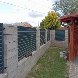 KRIS-STAL - Nowoczesne ogrodzenie z szarych, betonowych bloczków i poziomych, ciemnych paneli metalowych. Na murku leży szlifierka kątowa. Tło: trawnik, budynek z czerwoną dachówką.