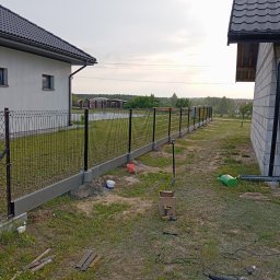 KRIS-STAL - Częściowy widok z poziomu gruntu na montaż ogrodzenia panelowego z podmurówką w Rakszawie. Widoczne narzędzia i elementy konstrukcyjne.