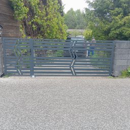 KRIS-STAL - Nowoczesna, szara brama metalowa z poziomymi panelami i geometrycznym wzorem, osadzona między słupkami z szarego betonu. W tle widoczny dom i zieleń.