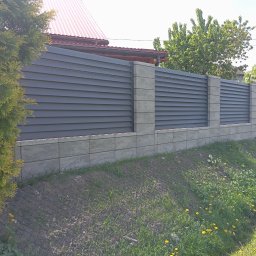 KRIS-STAL - Nowoczesne ogrodzenie z poziomych, szarych paneli i betonowych słupków, otoczone zielenią. Widoczny fragment domu i samochodu w tle. Słoneczny dzień.