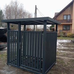 KRIS-STAL - Metalowa zabudowa śmietnikowa z pionowymi panelami w kolorze grafitowym, zamykana na zamek, na betonowej podstawie. W tle dom jednorodzinny i fragment otwartego bagażnika auta.