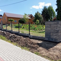 KRIS-STAL - Budowa ogrodzenia z bloczków betonowych w ogrodzie. Widoczny fragment fundamentu i słupka z bloczków. W tle dom z brązowym dachem i zieleń ogrodu.