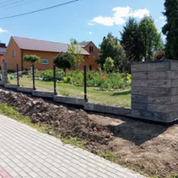 Budowa ogrodzenia z bloczków betonowych w ogrodzie. Widoczny fragment fundamentu i słupka z bloczków. W tle dom z brązowym dachem i zieleń ogrodu.
