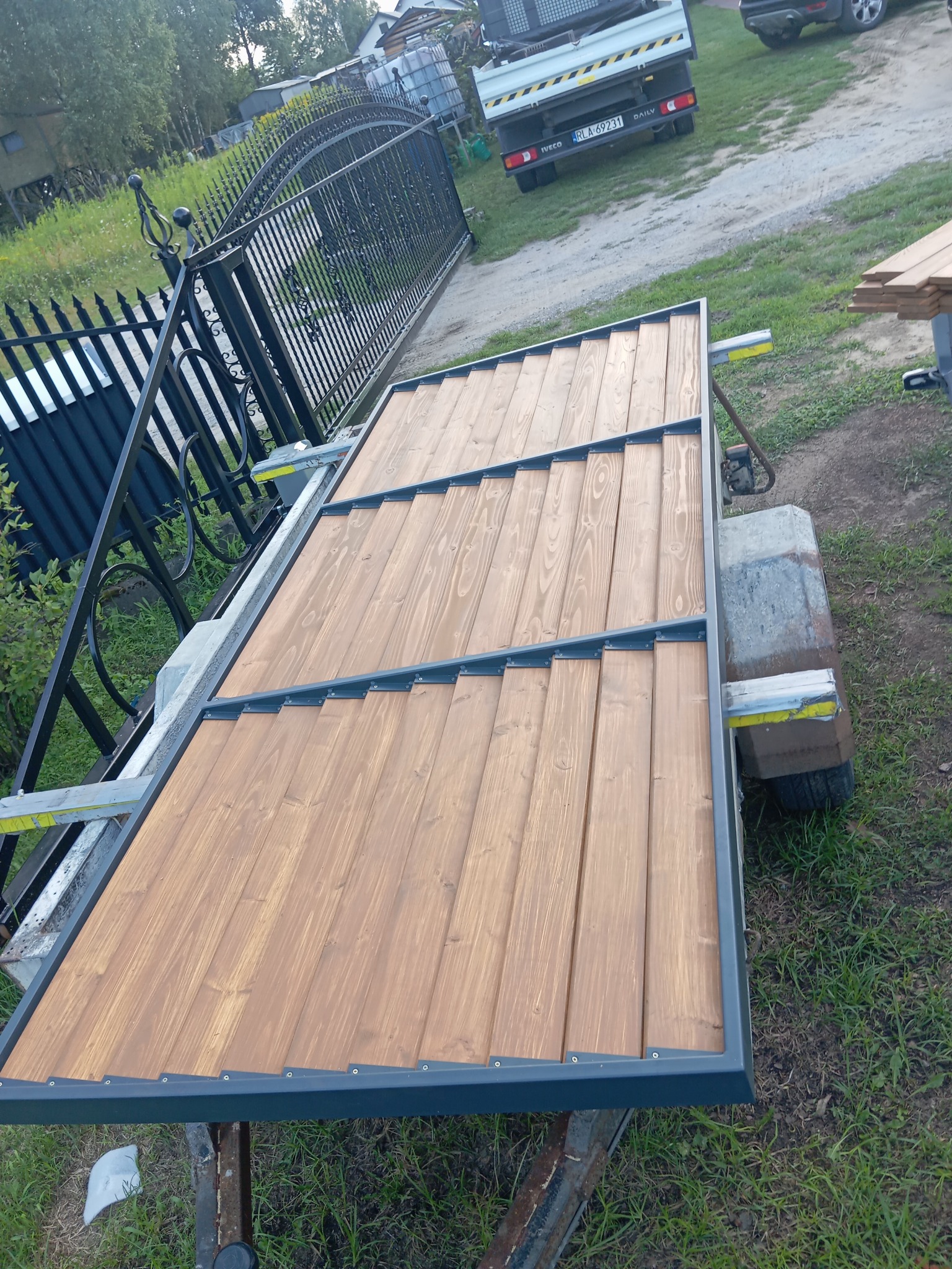 Brama z drewnianymi panelami w stalowej ramie, częściowo zamontowana, widoczny fragment ogrodzenia i samochód dostawczy w tle.