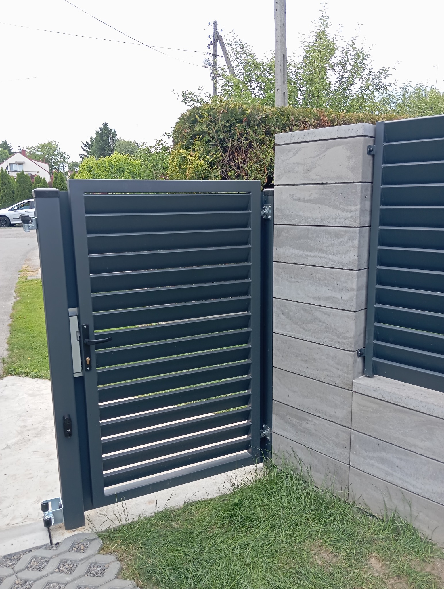 Nowoczesna, otwarta brama lamelowa w grafitowym kolorze, zamontowana na betonowym słupku z widocznymi poziomymi warstwami. Brama częściowo otwarta, z widocznym zamkiem.