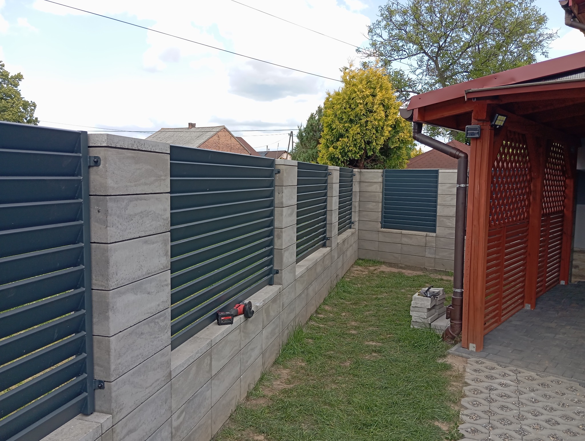 Nowoczesne ogrodzenie z szarych, betonowych bloczków i poziomych, ciemnych paneli metalowych. Na murku leży szlifierka kątowa. Tło: trawnik, budynek z czerwoną dachówką.