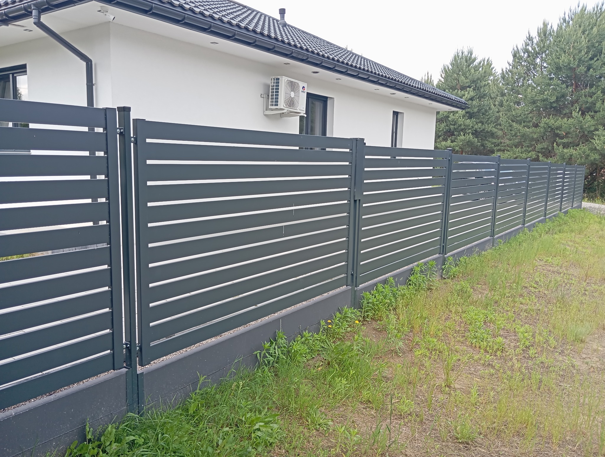 Nowoczesne ogrodzenie metalowe z poziomych paneli w kolorze grafitowym, otaczające dom z klimatyzacją. Widok z perspektywy trawnika.