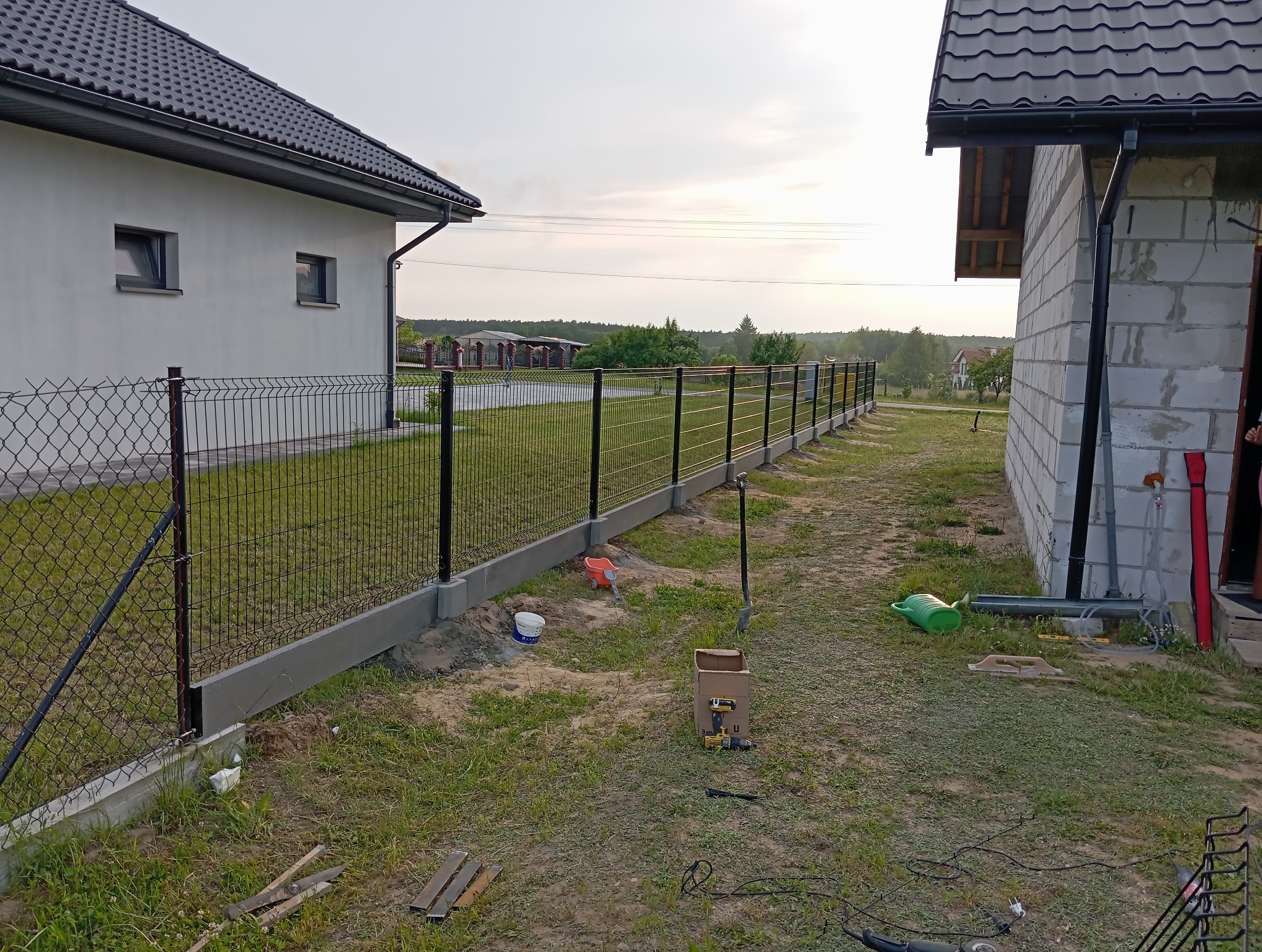 Częściowy widok z poziomu gruntu na montaż ogrodzenia panelowego z podmurówką w Rakszawie. Widoczne narzędzia i elementy konstrukcyjne.