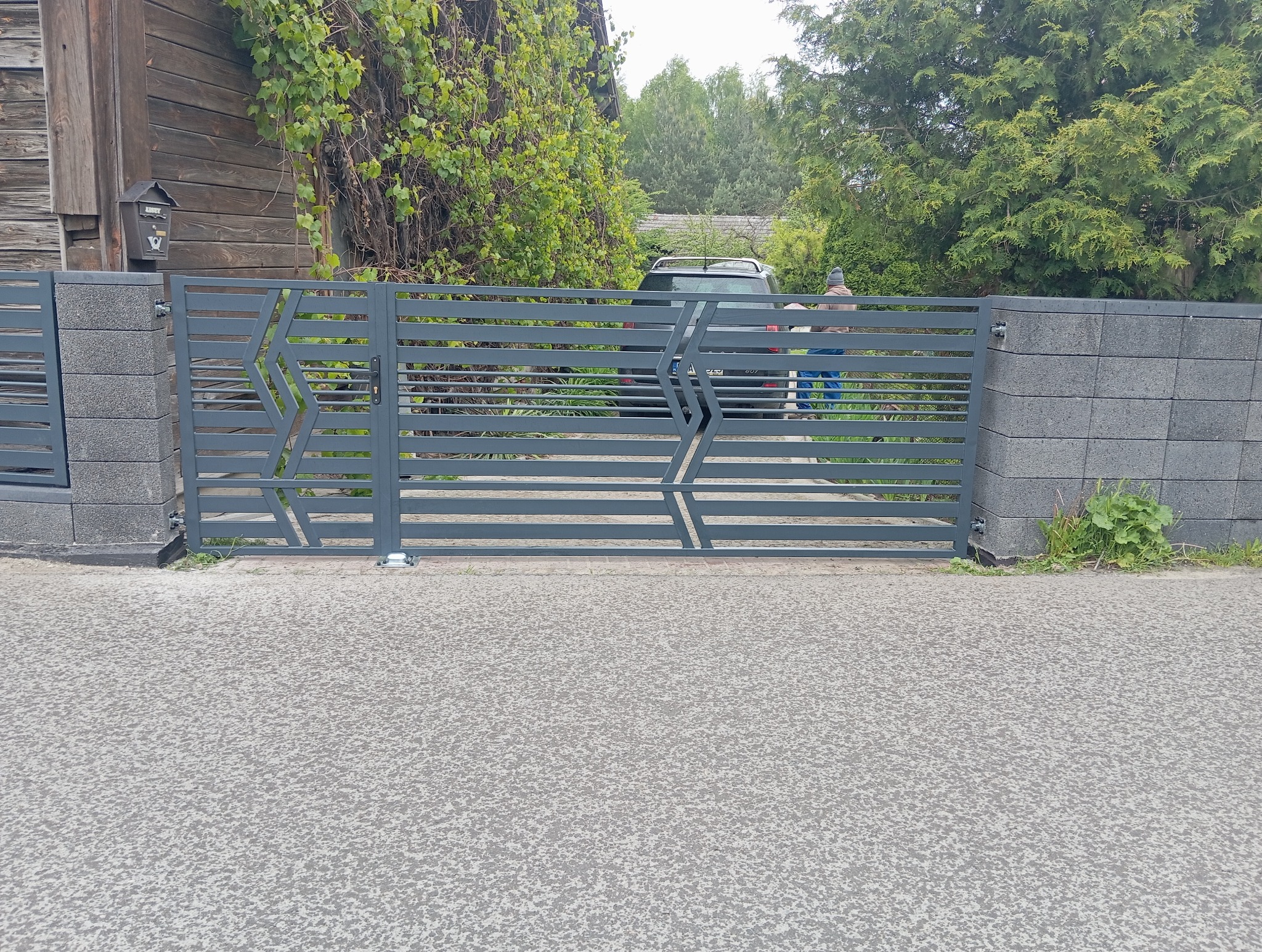 Nowoczesna, szara brama metalowa z poziomymi panelami i geometrycznym wzorem, osadzona między słupkami z szarego betonu. W tle widoczny dom i zieleń.