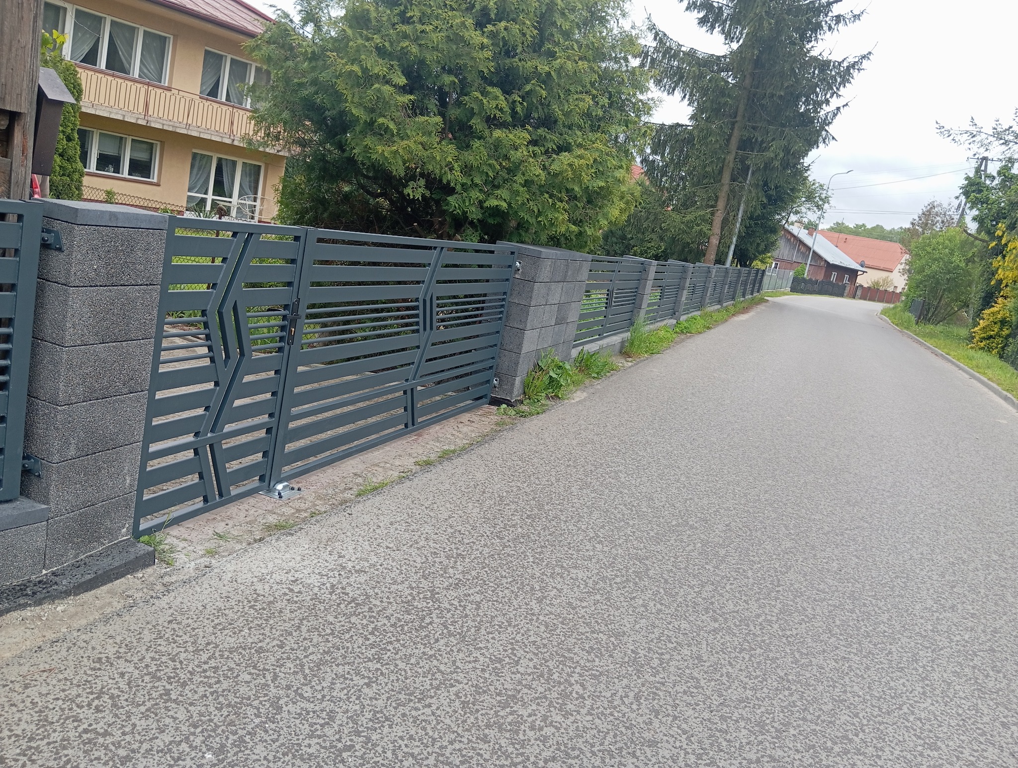 Nowoczesne ogrodzenie panelowe z szarymi, betonowymi słupkami i metalowymi przęsłami o geometrycznym wzorze, widoczne z perspektywy ulicy.