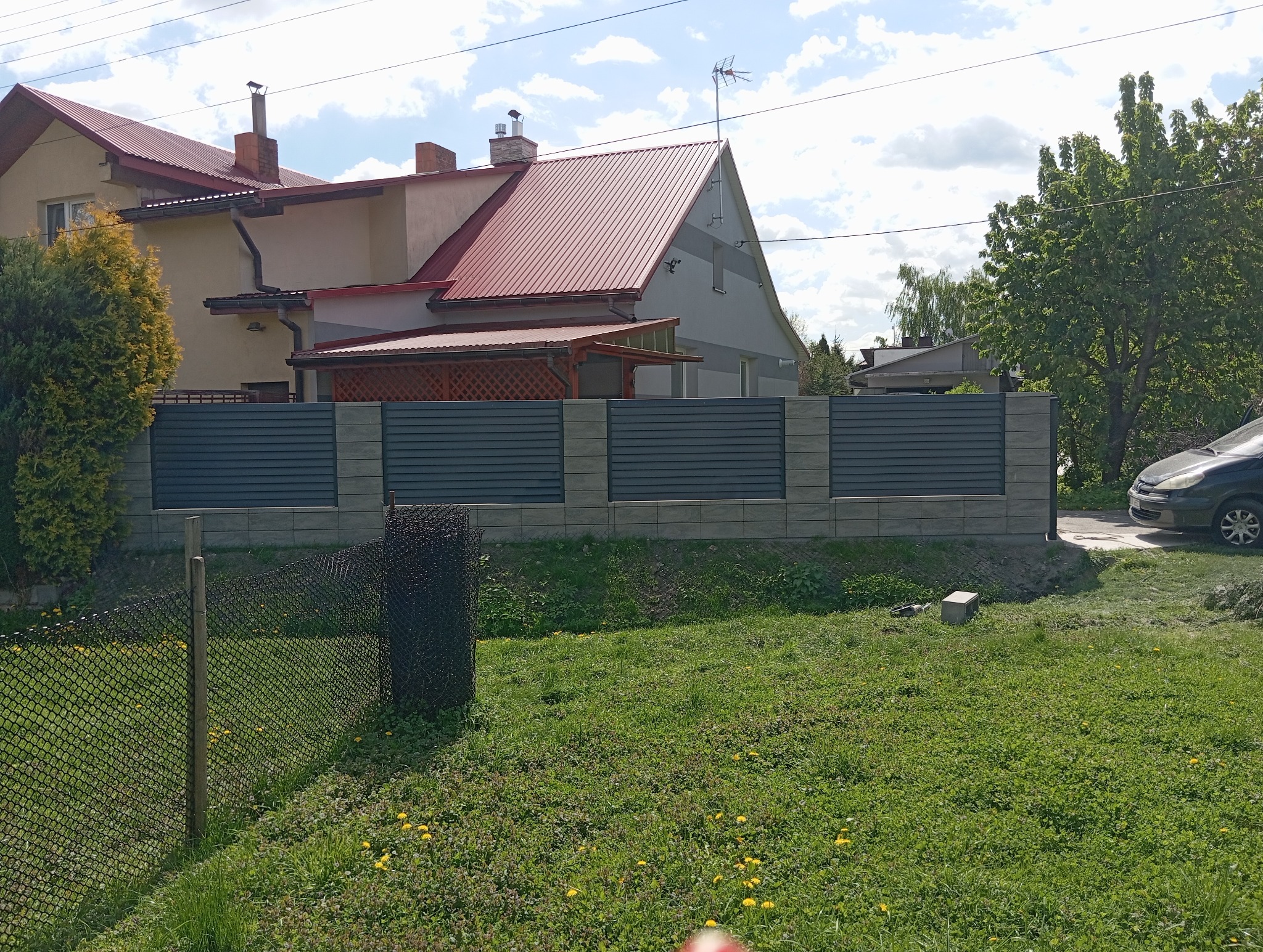 Nowoczesne ogrodzenie z poziomych paneli w kolorze grafitowym, osadzone na betonowych słupkach, na tle domu z czerwoną dachówką i bujnej zieleni.