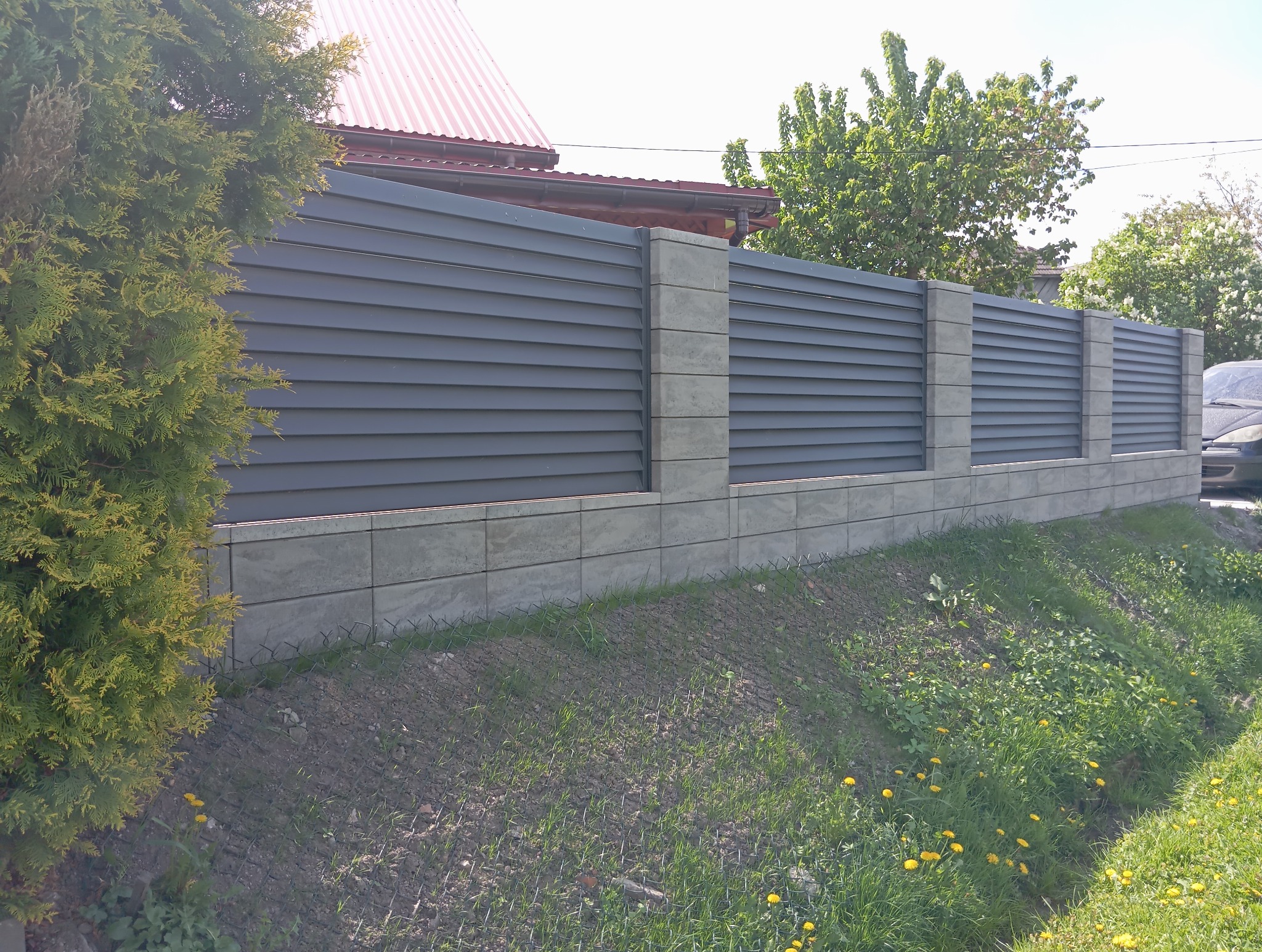 Nowoczesne ogrodzenie z poziomych, szarych paneli i betonowych słupków, otoczone zielenią. Widoczny fragment domu i samochodu w tle. Słoneczny dzień.