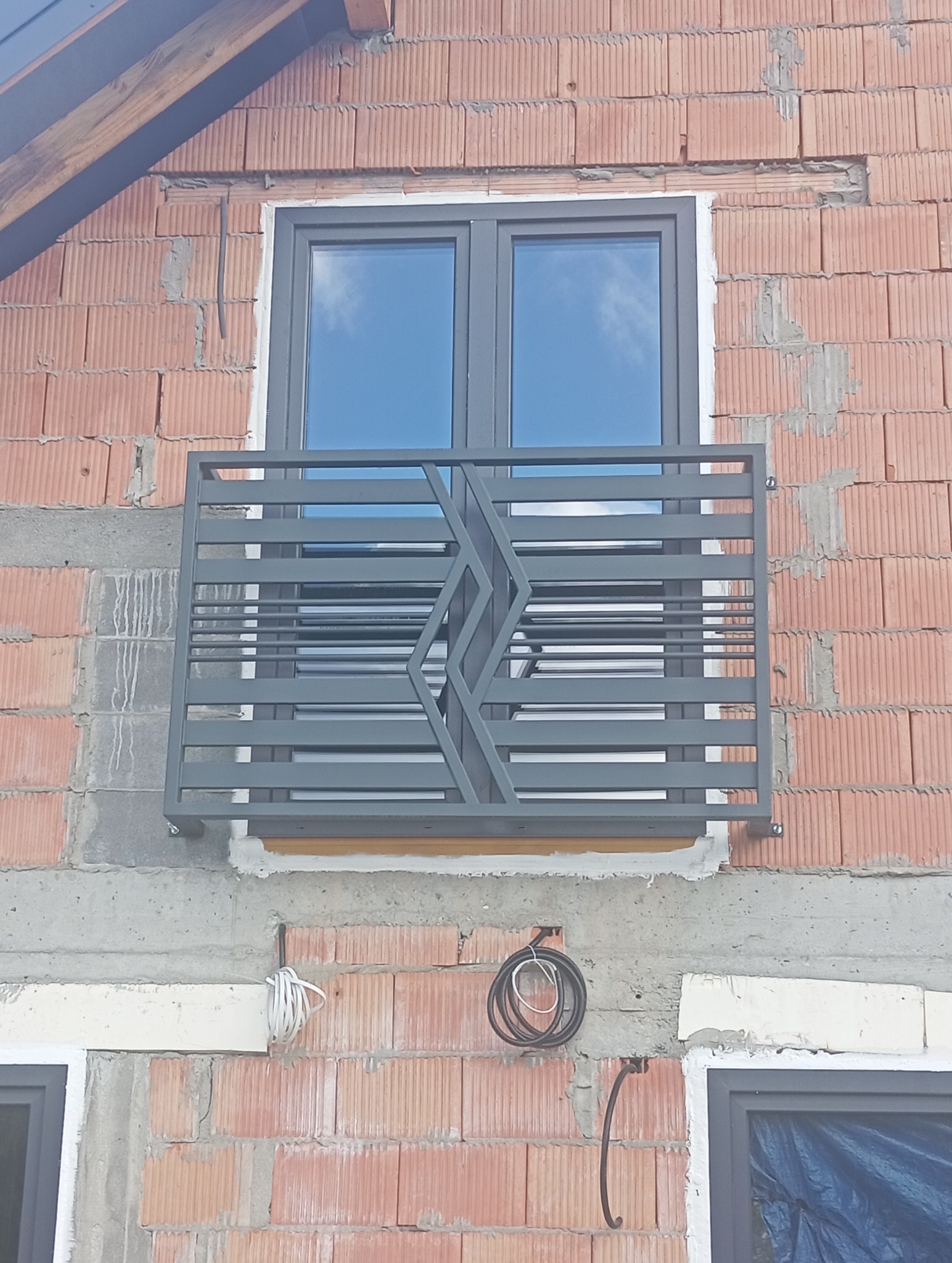 Szara, metalowa balustrada okienna z geometrycznym wzorem na tle okna i surowej ściany z cegły. Widoczne przewody elektryczne. Balustrada zabezpiecza okno na zewnątrz.