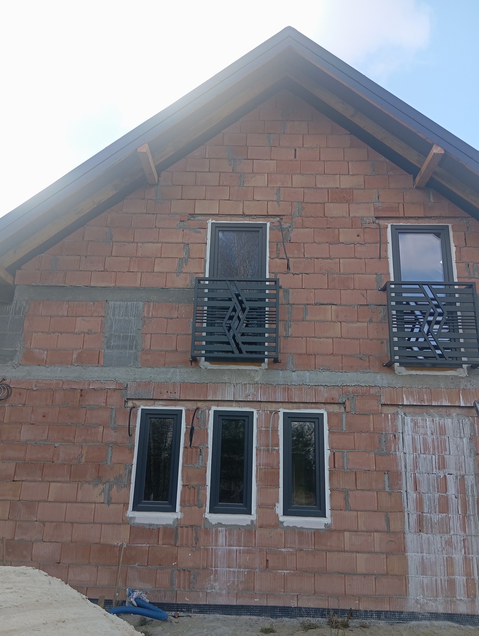 Surowa elewacja budynku z zamontowanymi, nowoczesnymi balustradami balkonowymi o geometrycznym wzorze. Widoczne okna i początek prac wykończeniowych.