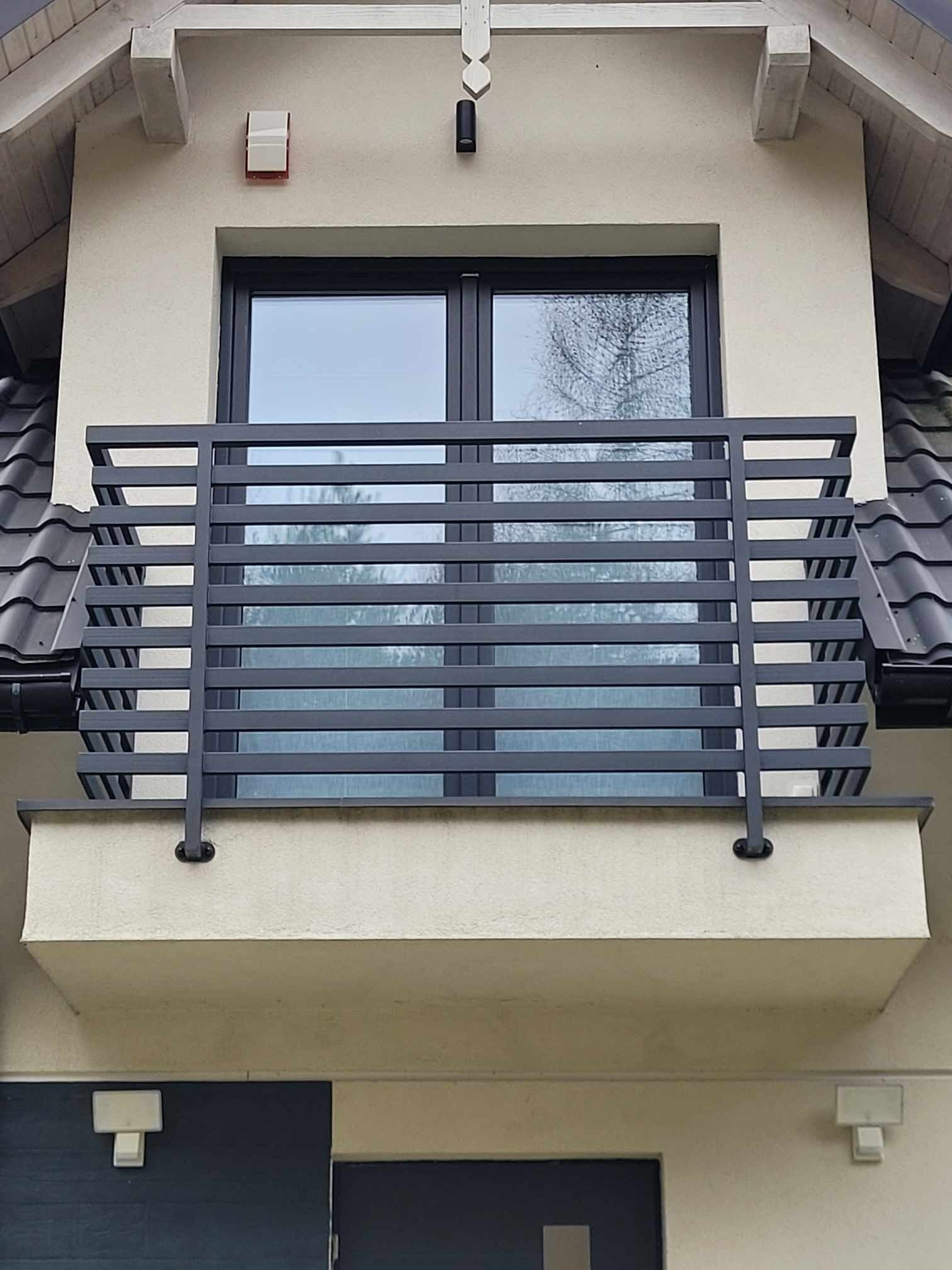 Szara, pozioma balustrada balkonowa z metalowych profili, zamontowana na elewacji budynku z oknem. Widoczne fragmenty dachu z czarną dachówką.