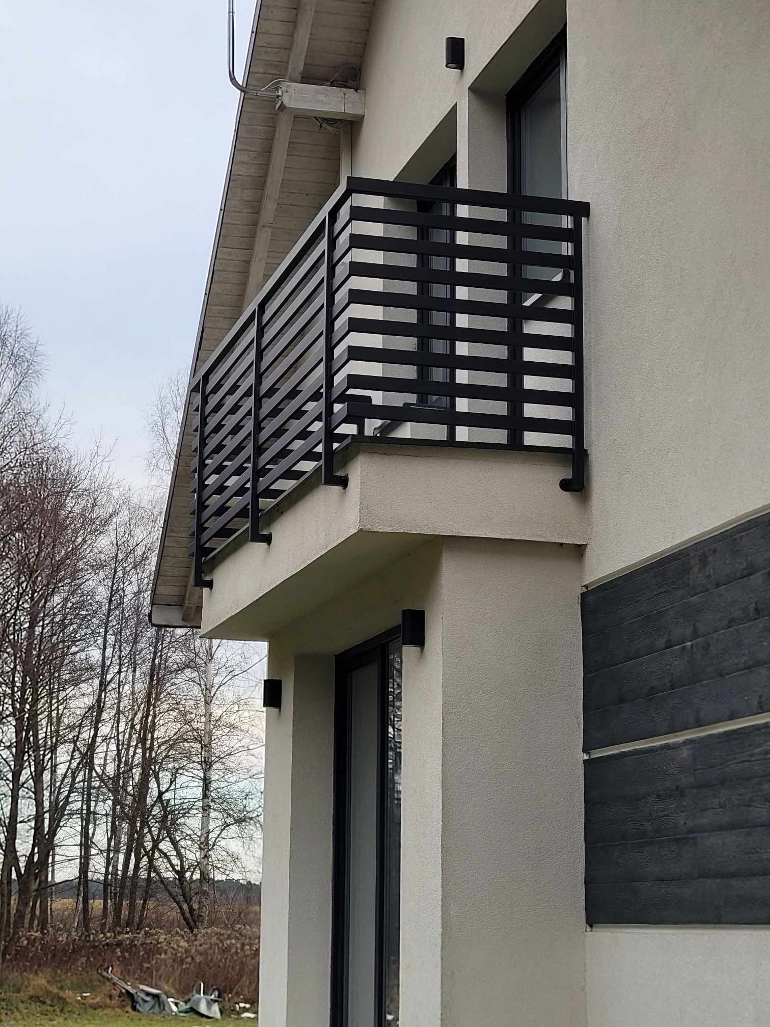 Nowoczesna, czarna balustrada balkonowa z poziomymi szczebelkami, zamontowana przy jasnym budynku z elementami ciemnego drewna. Widok z dołu na balkon.