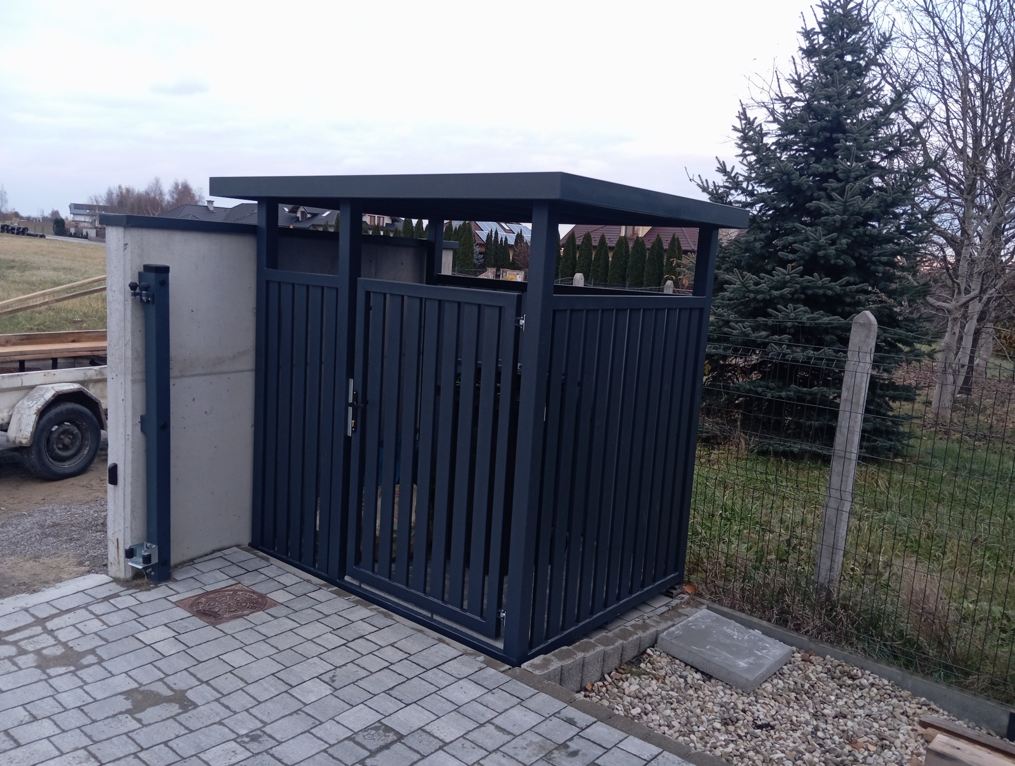 Nowoczesna, grafitowa obudowa śmietnikowa z metalowymi lamelami i płaskim dachem, zamykana na zamek, na tle kostki brukowej i zieleni ogrodu.