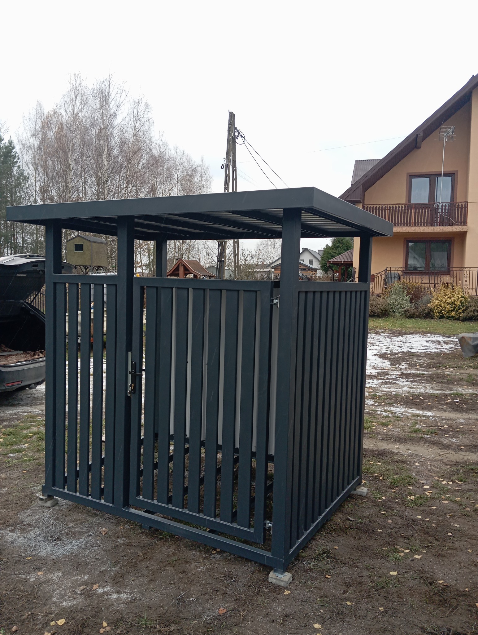 Metalowa zabudowa śmietnikowa z pionowymi panelami w kolorze grafitowym, zamykana na zamek, na betonowej podstawie. W tle dom jednorodzinny i fragment otwartego bagażnika auta.