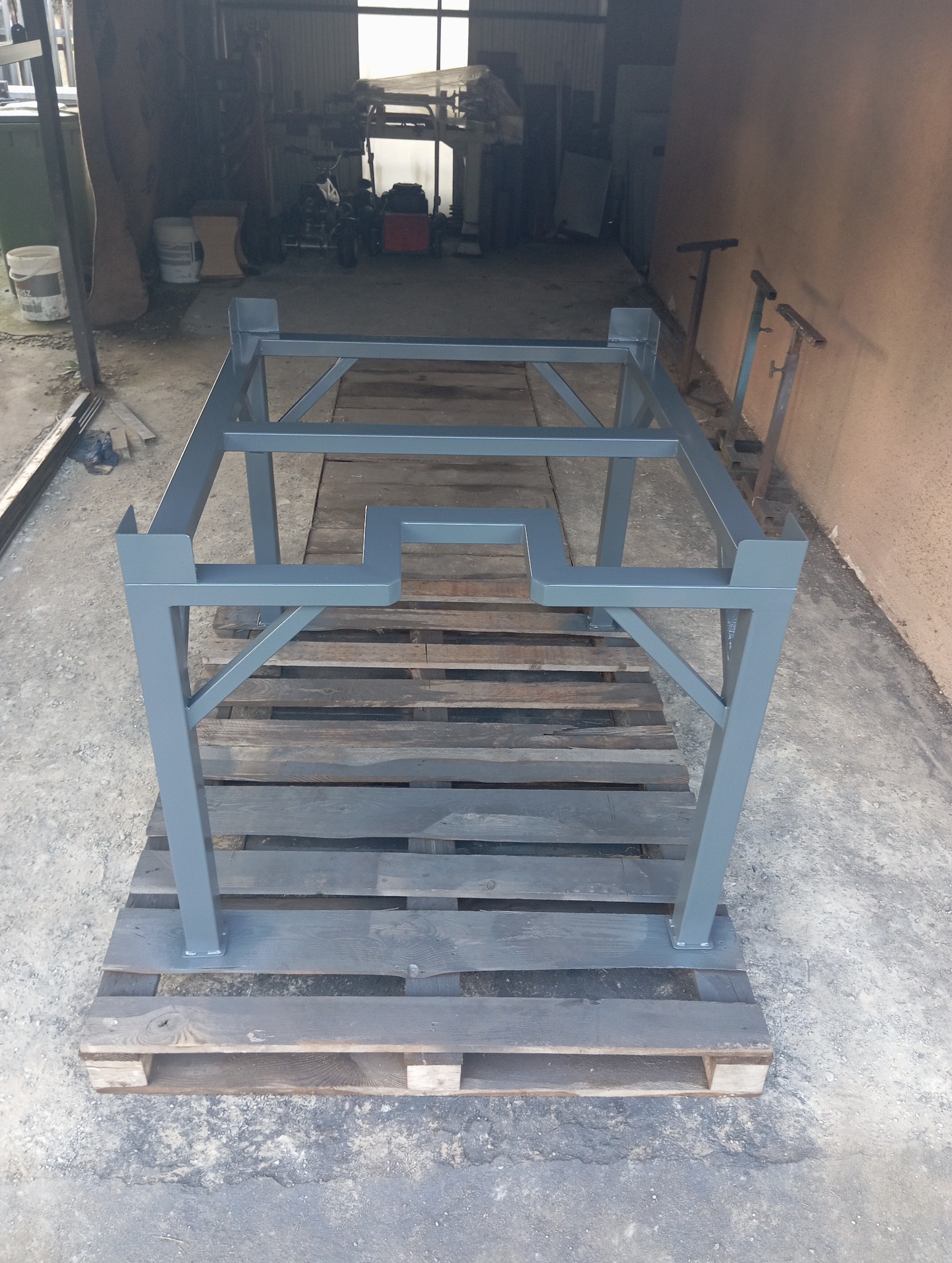 Szara, metalowa konstrukcja spawana na palecie w warsztacie. Element z profili zamkniętych, pomalowany. Widoczne spawy i fragmenty wyposażenia warsztatu w tle.