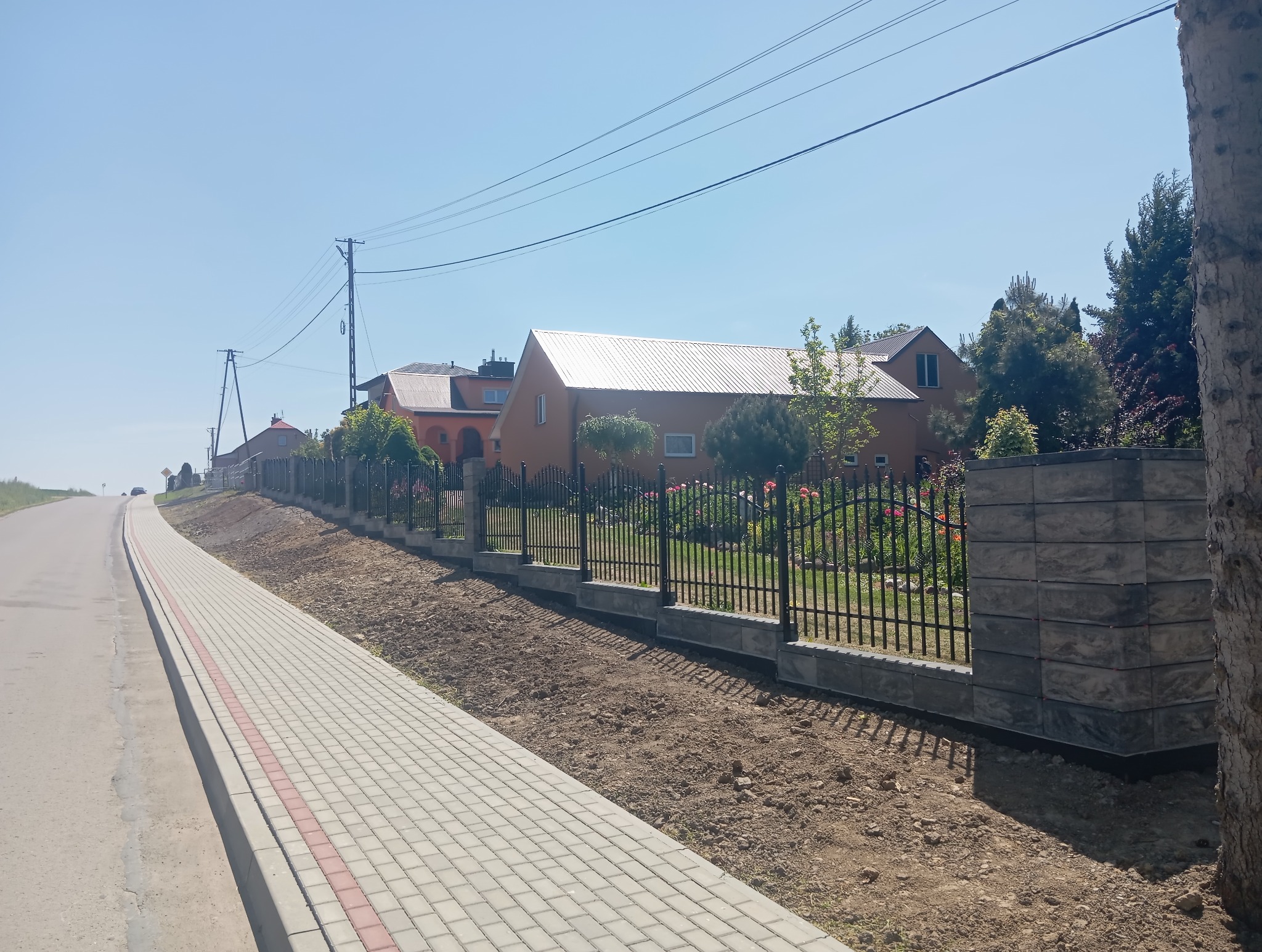 Nowoczesne ogrodzenie metalowe z podmurówką z szarej kostki, oddzielające posesję od chodnika i ulicy. W tle domy jednorodzinne i błękitne niebo.