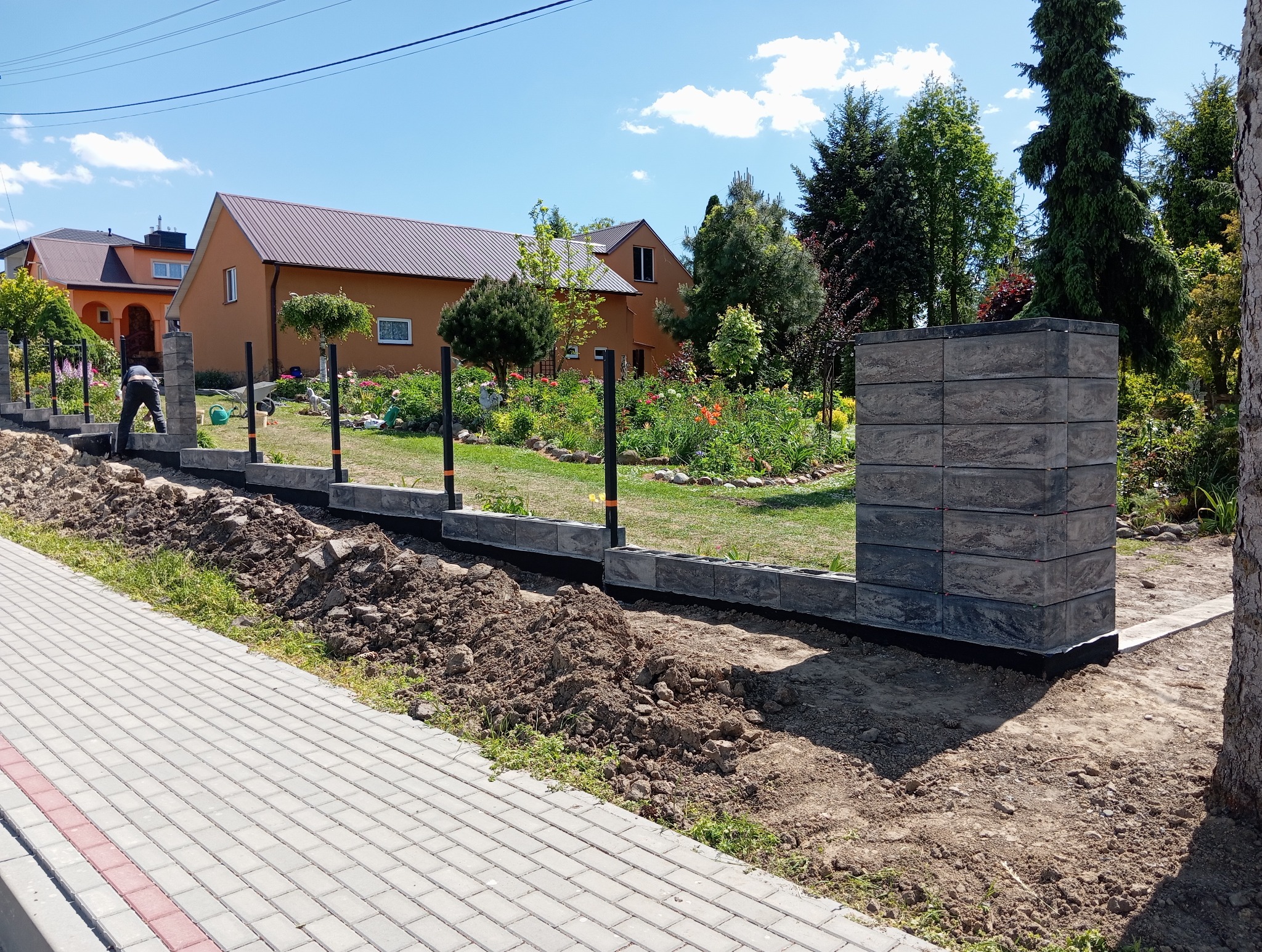 Budowa ogrodzenia z bloczków betonowych w ogrodzie. Widoczny fragment fundamentu i słupka z bloczków. W tle dom z brązowym dachem i zieleń ogrodu.