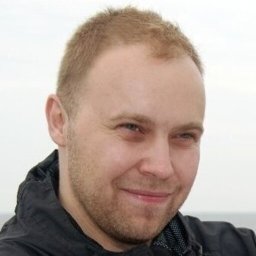 Akto Usługi Internetowe - Portret mężczyzny z krótkimi włosami i lekkim zarostem, ubranego w ciemną kurtkę, na rozmytym tle. Uśmiechnięty, patrzy w obiektyw.