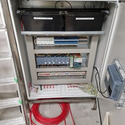 Wattman - Otwarta szafa sterownicza z akumulatorami ALARMTEC, bezpiecznikami, modułami sterującymi i okablowaniem. Widoczna drabina i zwój czerwonego kabla na podłodze.