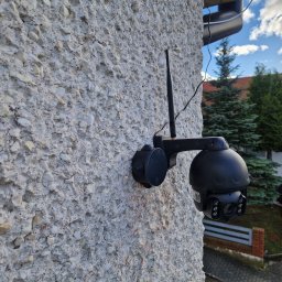 Wattman - Zewnętrzna kamera monitoringu PTZ z anteną, zamontowana na szorstkiej, jasnej elewacji budynku. Widoczne okablowanie i fragment otoczenia z zielenią i samochodem.