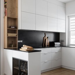 Liczy się wnętrze Justyna Kućmierz - Nowoczesna kuchnia z białymi szafkami, czarnym blatem i drewnianymi akcentami. Na blacie leżą bagietki. Widoczne szklanki w szafce pod blatem i deski do krojenia.