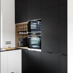 Liczy się wnętrze Justyna Kućmierz - Nowoczesna kuchnia z czarnymi, matowymi frontami szafek, drewnianym regałem na wino i sprzętem AGD w zabudowie, podkreślona jasną podłogą i blatem.