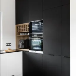 Nowoczesna kuchnia z czarnymi, matowymi frontami szafek, drewnianym regałem na wino i sprzętem AGD w zabudowie, podkreślona jasną podłogą i blatem.