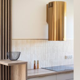 Liczy się wnętrze Justyna Kućmierz - Nowoczesna kuchnia z złotym okapem cylindrycznym, drewnianą ścianką z listew i blatem w kolorze drewna. Minimalistyczny design, płytki w odcieniach beżu.
