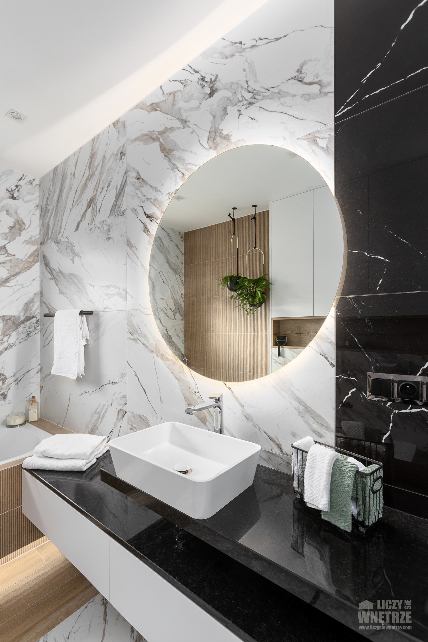 Elegancka łazienka z okrągłym lustrem podświetlanym LED, białą umywalką nablatową i czarnym blatem. Marmurowe płytki w odcieniach bieli i czerni.