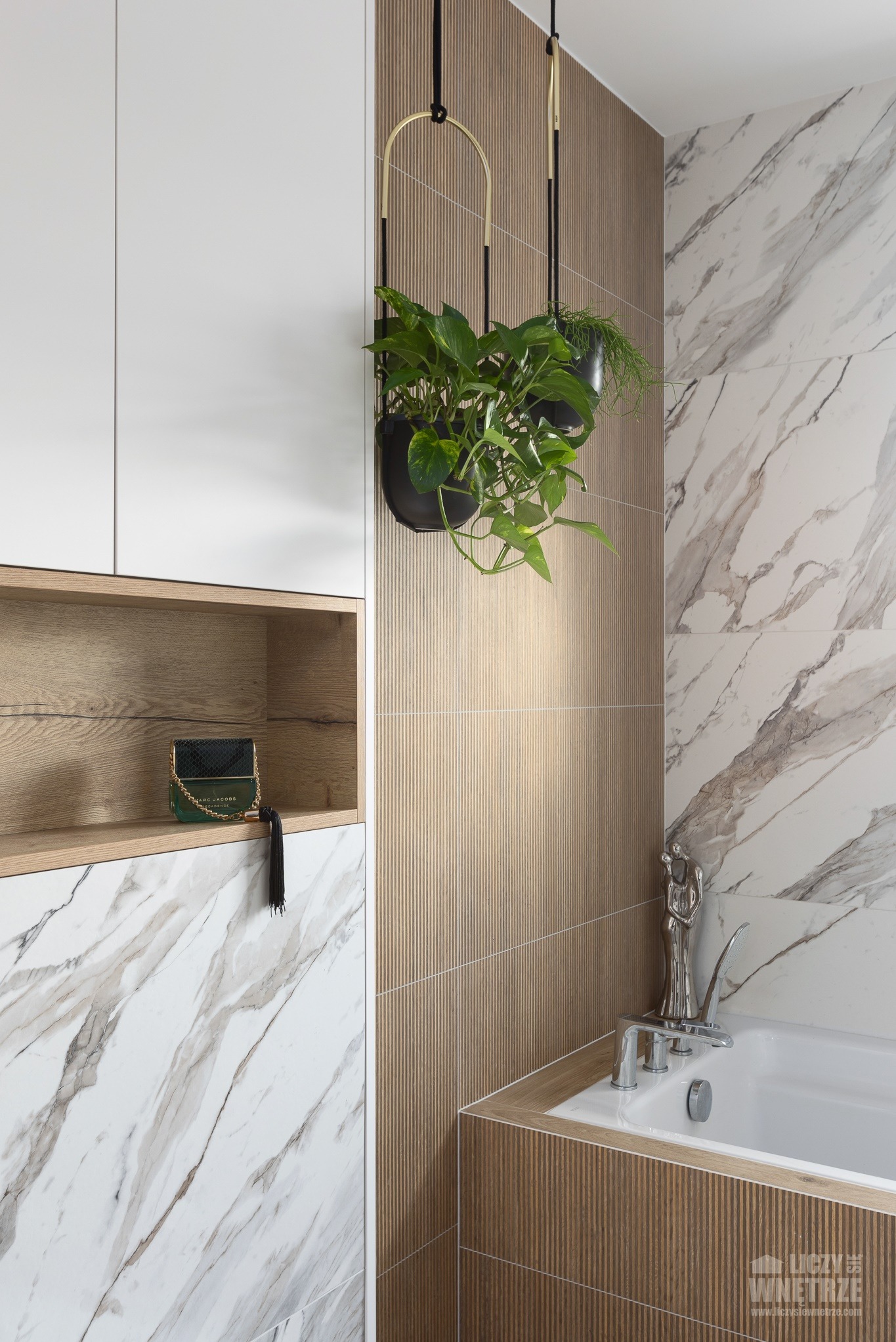 Nowoczesna łazienka z marmurowymi płytkami, drewnianymi akcentami i wiszącymi roślinami. Na półce torebka Marc Jacobs, a obok wanny srebrna figurka.