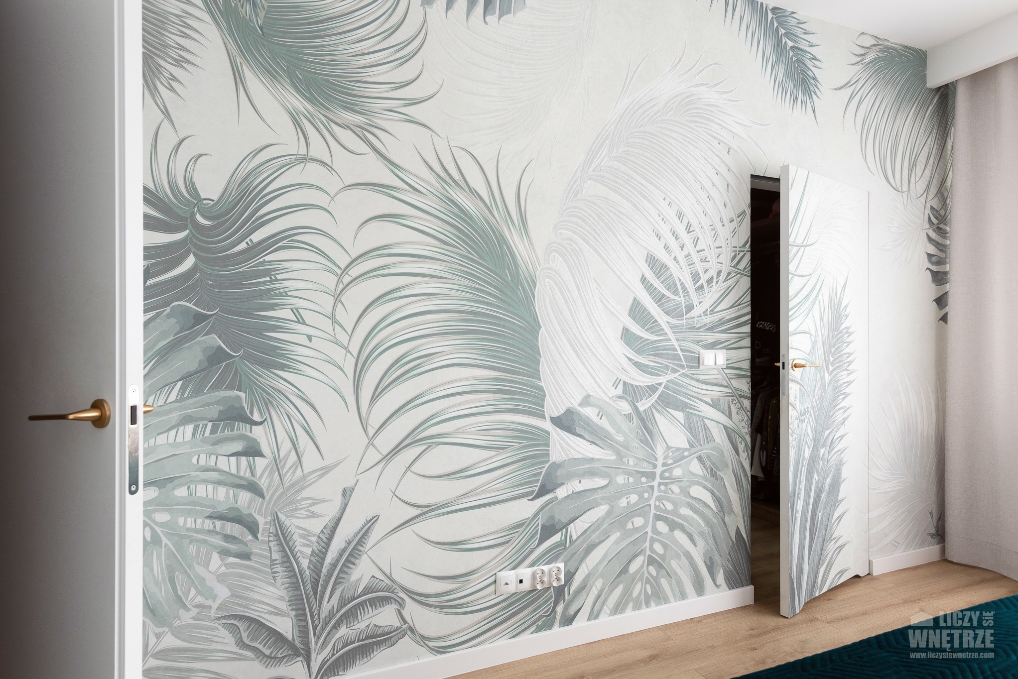 Pokój z tapetą w tropikalne liście, białe drzwi z złotymi klamkami, drewniana podłoga. Widok na otwartą garderobę i zasłony. Jasne, naturalne światło.