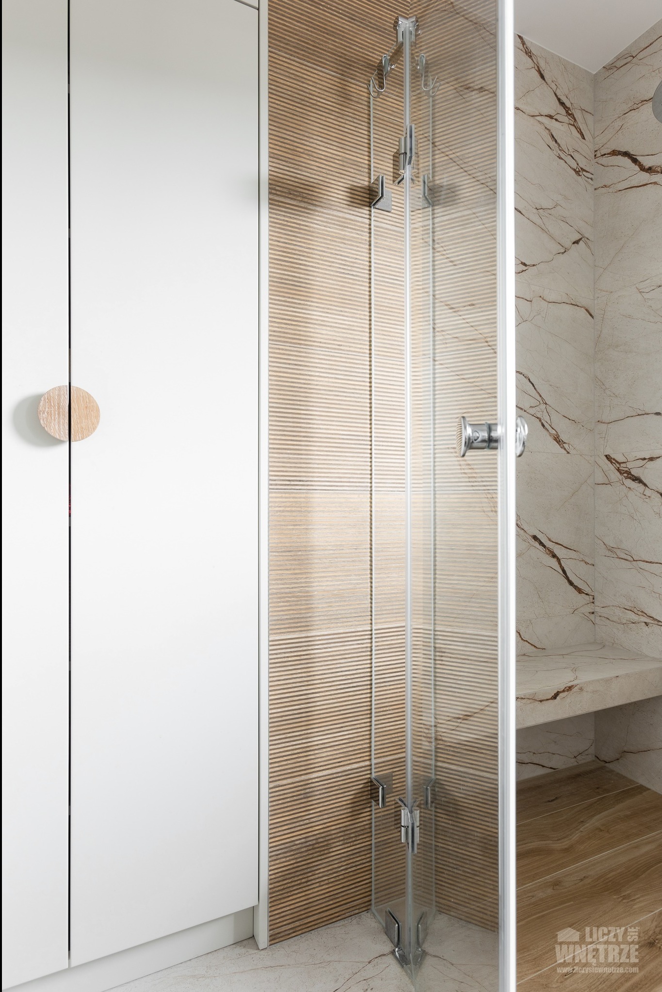Nowoczesna kabina prysznicowa z drewnianymi akcentami i marmurowym wykończeniem w łazience. Widoczna armatura i detale wykończeniowe.