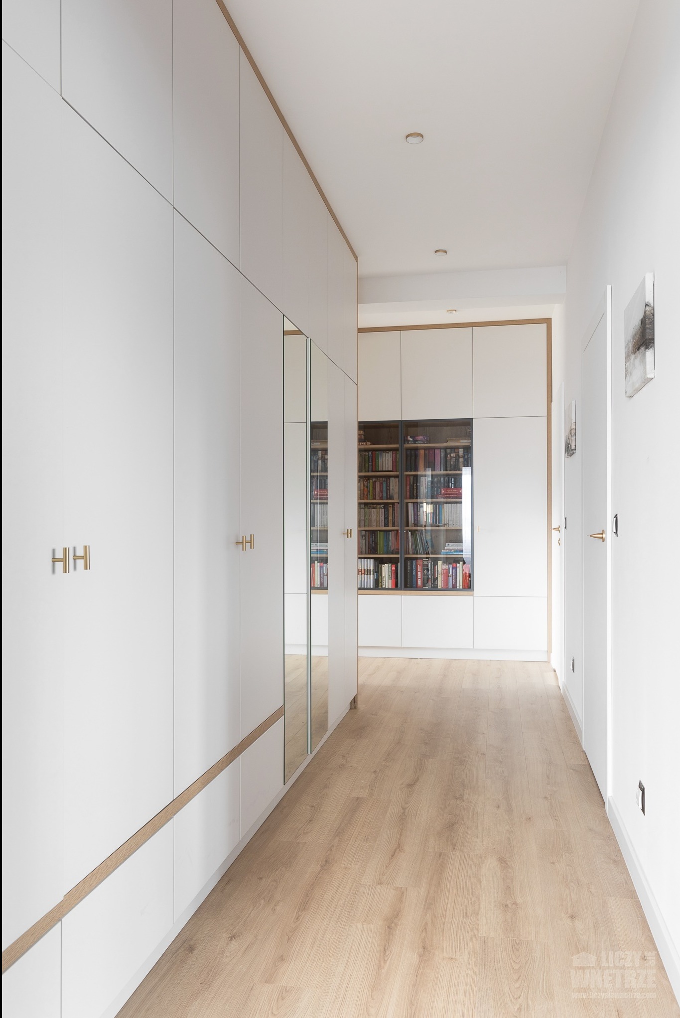 Białe szafy wnękowe z lustrem i drewnianymi akcentami w korytarzu. Widoczny regał z książkami w głębi. Jasna podłoga z paneli.