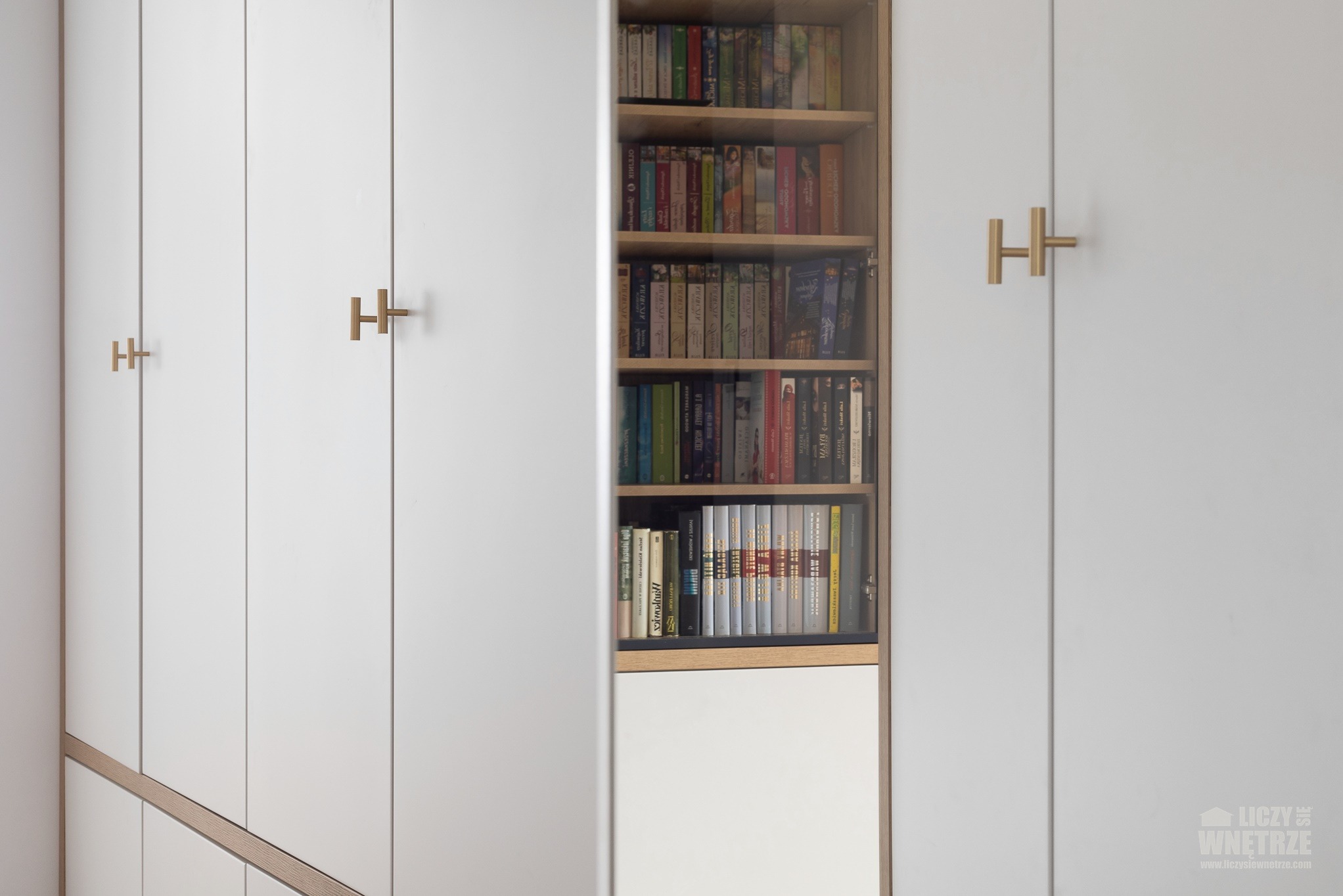 Biała zabudowa meblowa z półkami na książki, otwarte drzwi szafy eksponują regał. Minimalistyczne uchwyty w kształcie litery H. Jasne, stonowane wnętrze.