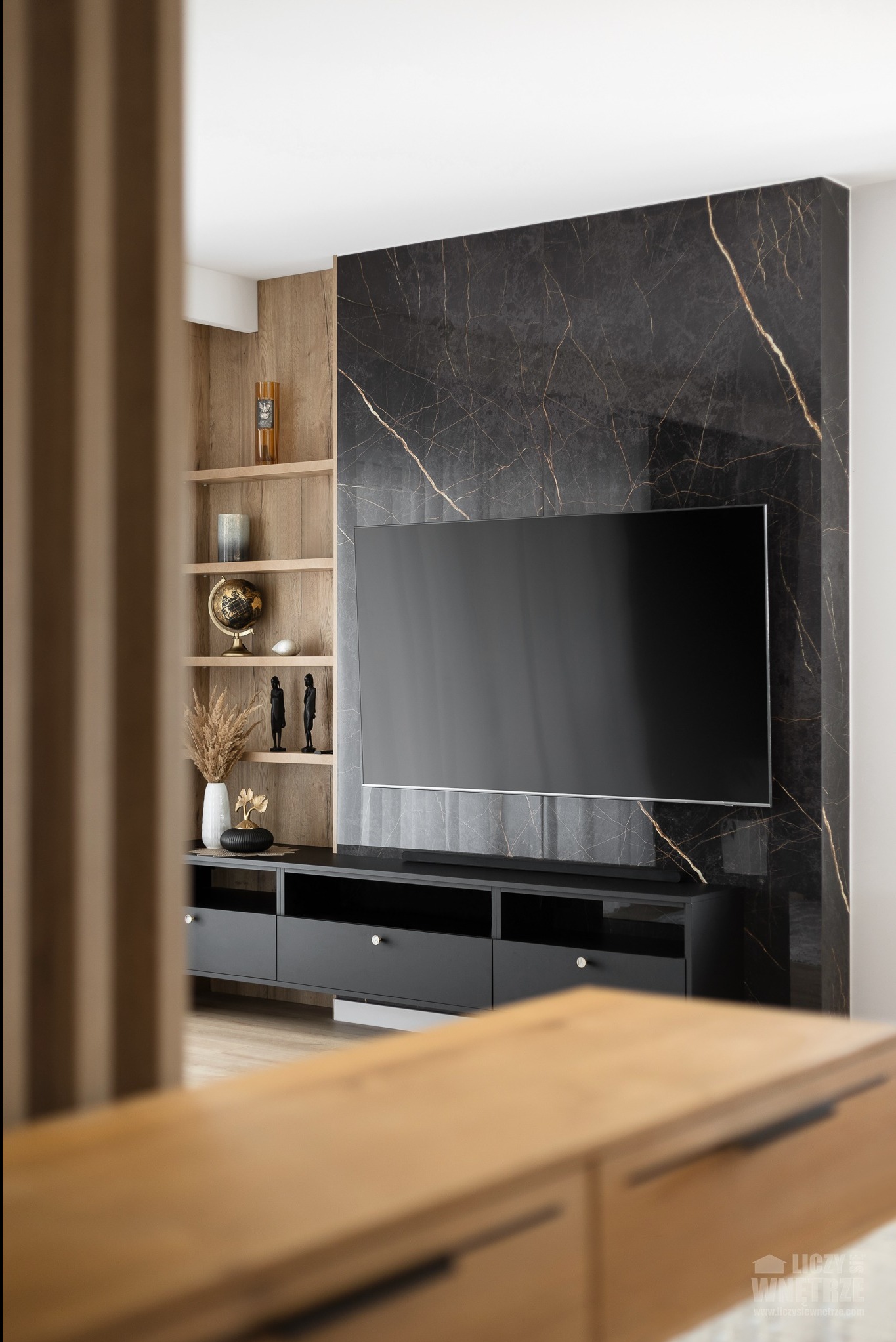 Stylowy salon z czarną ścianą z marmuru za telewizorem. Na półkach dekoracje: globus, wazon z trawą pampasową. Częściowo widoczny drewniany parawan.