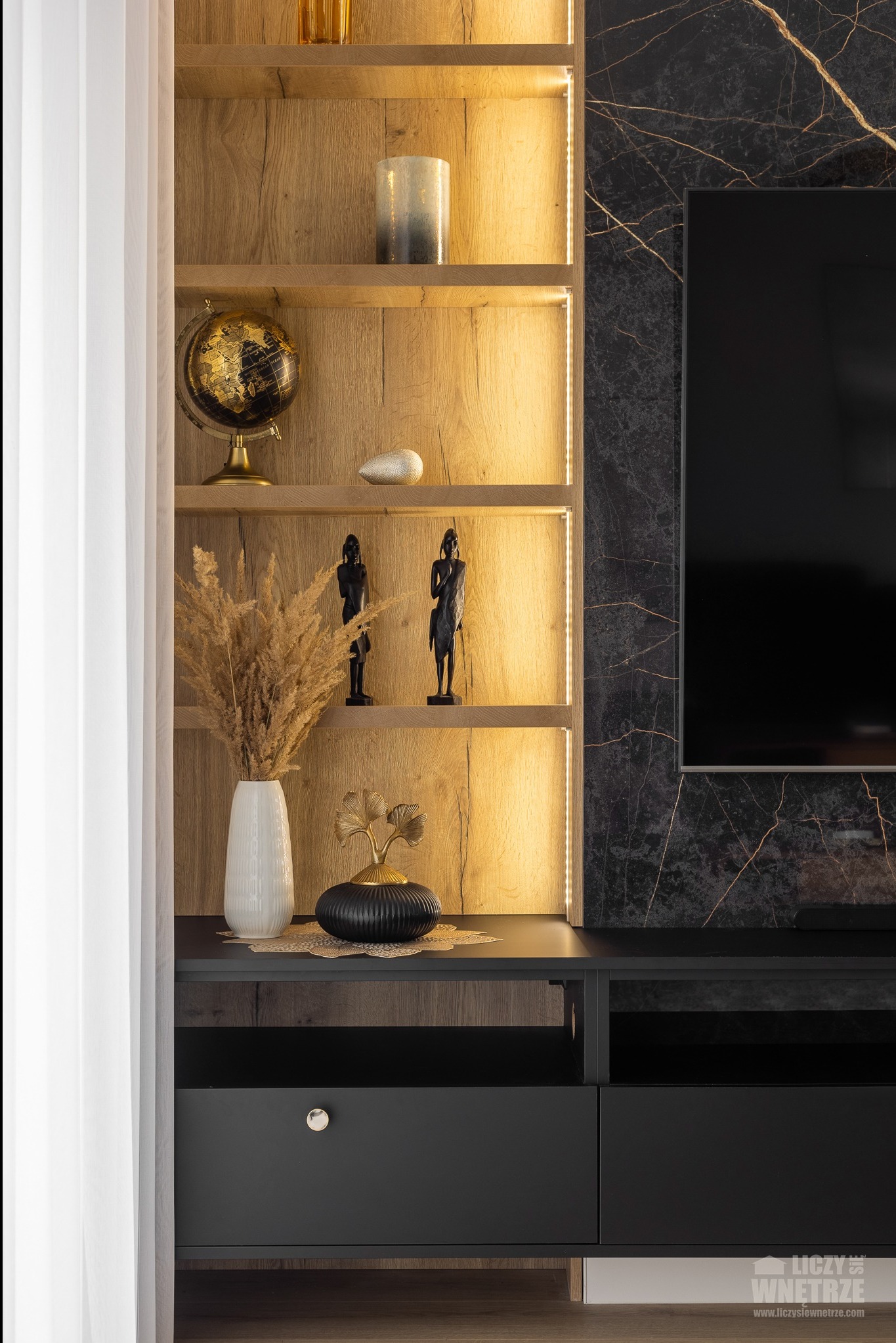 Stylowa aranżacja ściany TV z podświetlanymi półkami z dekoracjami: globus, figurki, wazon z suszem. Czarna szafka RTV z szufladą i marmurowa ściana.