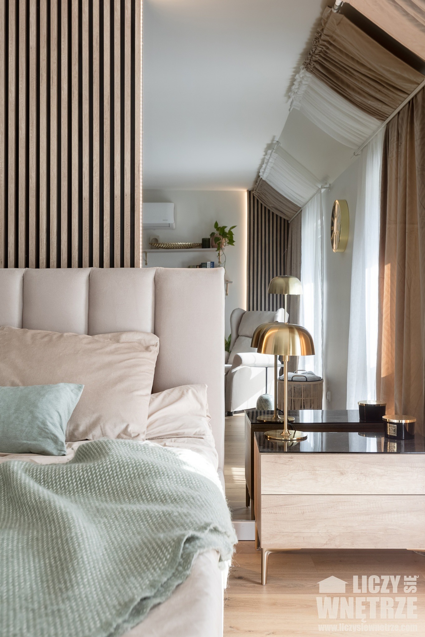 Elegancka sypialnia z beżowym łóżkiem, drewnianą ścianką działową i złotymi lampkami nocnymi. W tle widoczny salon z klimatyzacją i roślinami.