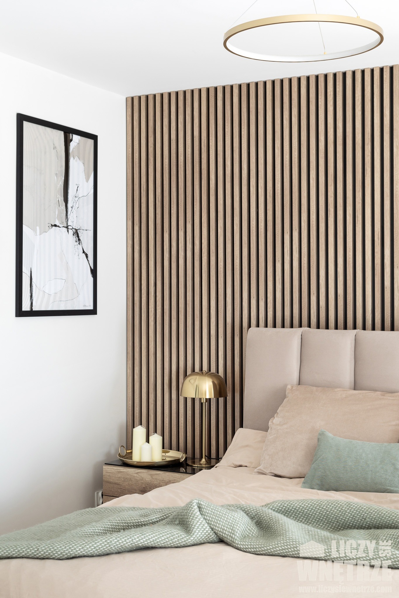 Nowoczesna sypialnia: drewniane lamele za łóżkiem, abstrakcyjny obraz w czarnej ramie, złota lampka nocna i pastelowa pościel z zielonym pledem. Subtelne oświetlenie sufitowe.