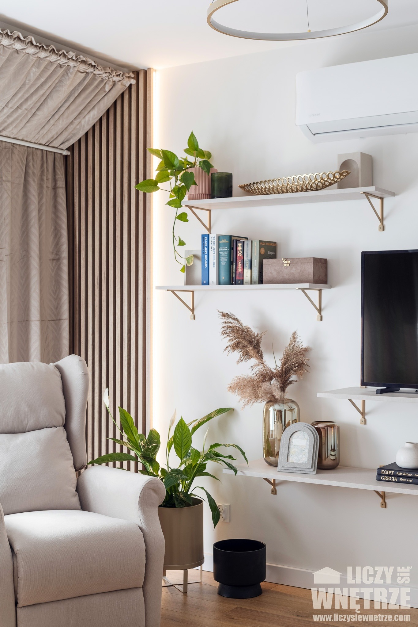 Jasny salon z beżowym fotelem, drewnianymi lamelami i półkami z dekoracjami. Na ścianie klimatyzator. Elementy wystroju wnętrza tworzą przytulną atmosferę.