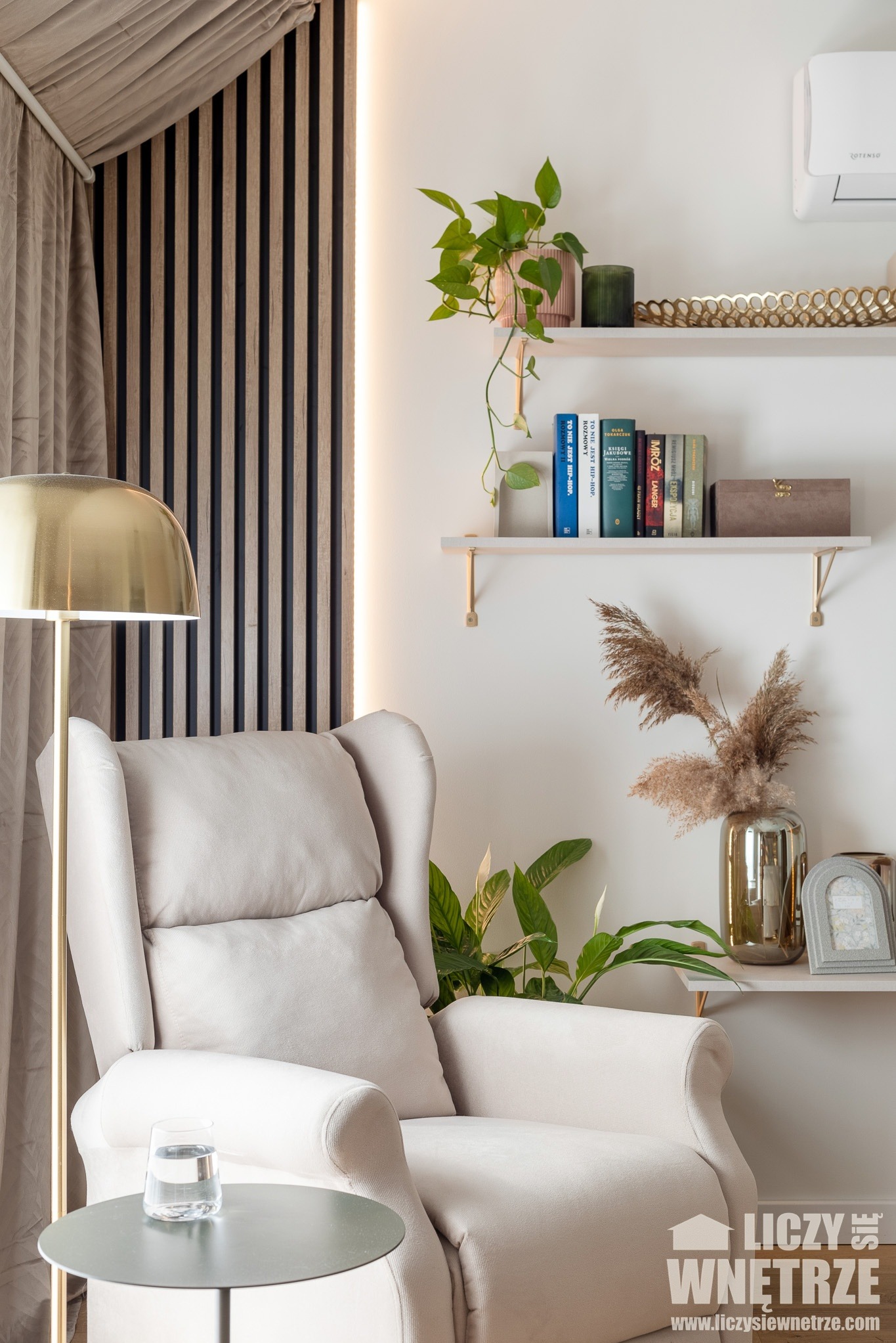 Beżowy fotel uszak z lampą i stolikiem w jasnym wnętrzu z drewnianą ścianą i półkami z dekoracjami. Minimalistyczny styl.